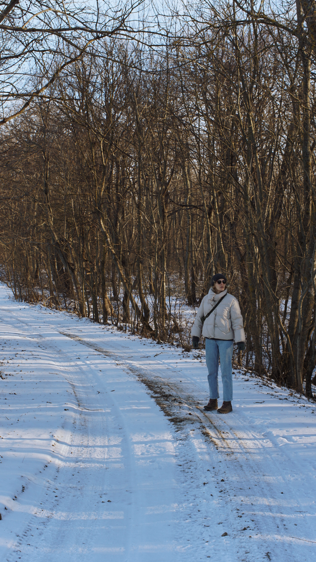 A winter hiker in a light puffer jacket walks along a snow-covered trail lined with bare trees.