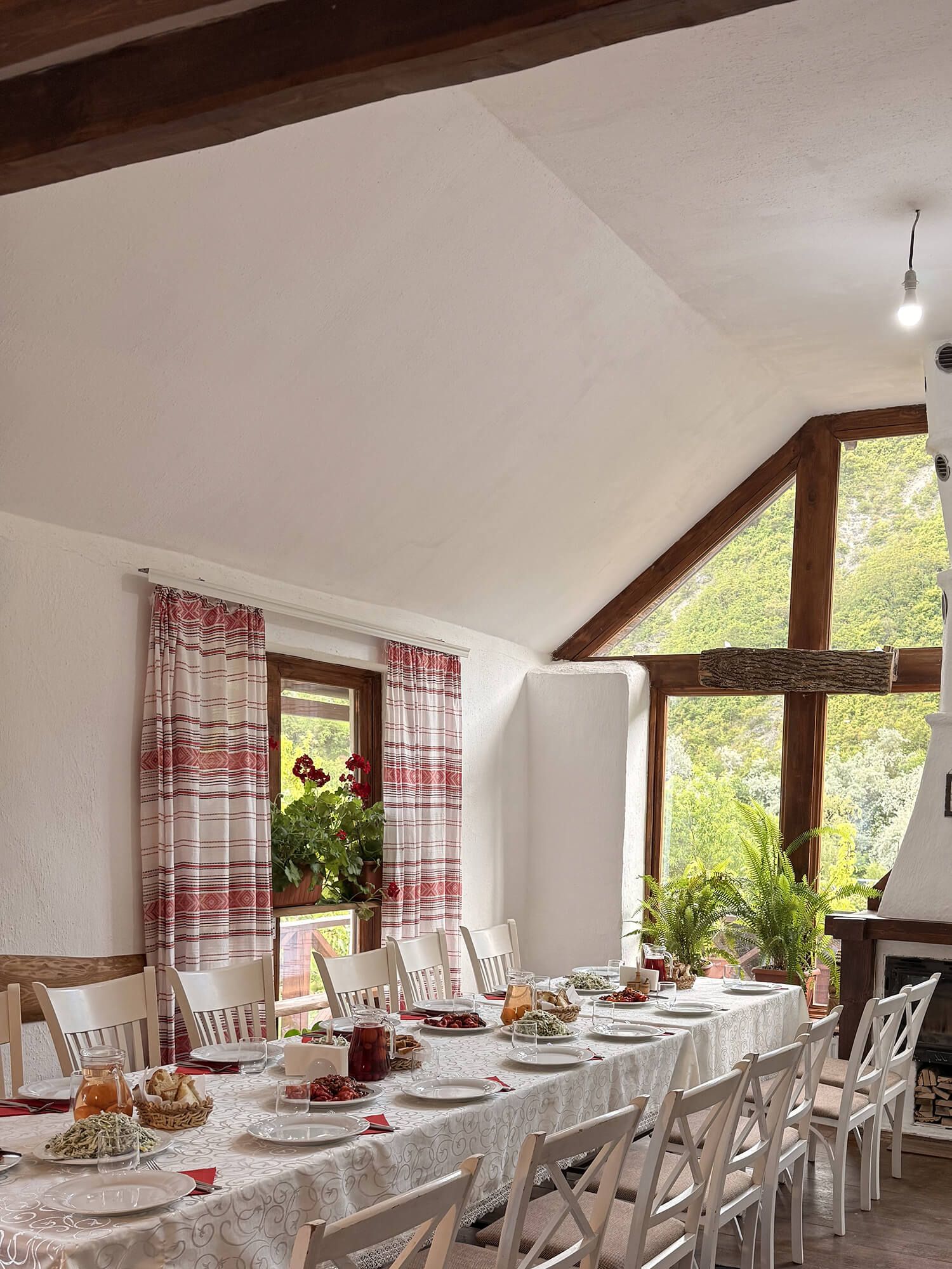 Rustic dining room with long white table set for guests, exposed wood beams, plaid curtains, and large windows overlooking greenery.