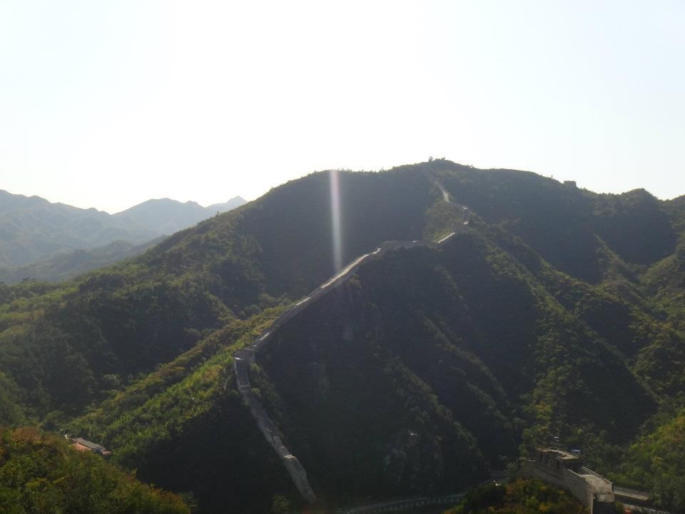 Sunlit mountain landscape showing winding paths along green ridges with a distant tower visible.