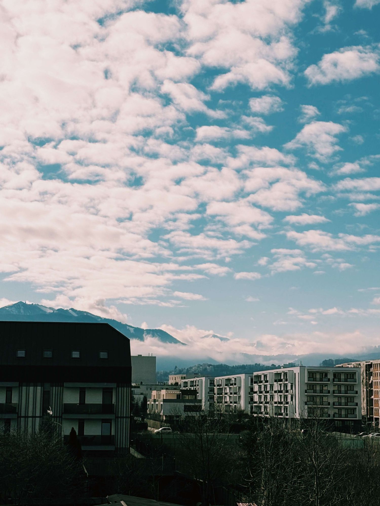 Modern residential buildings line a street against a bright blue sky with scattered white clouds and mountain backdrop.