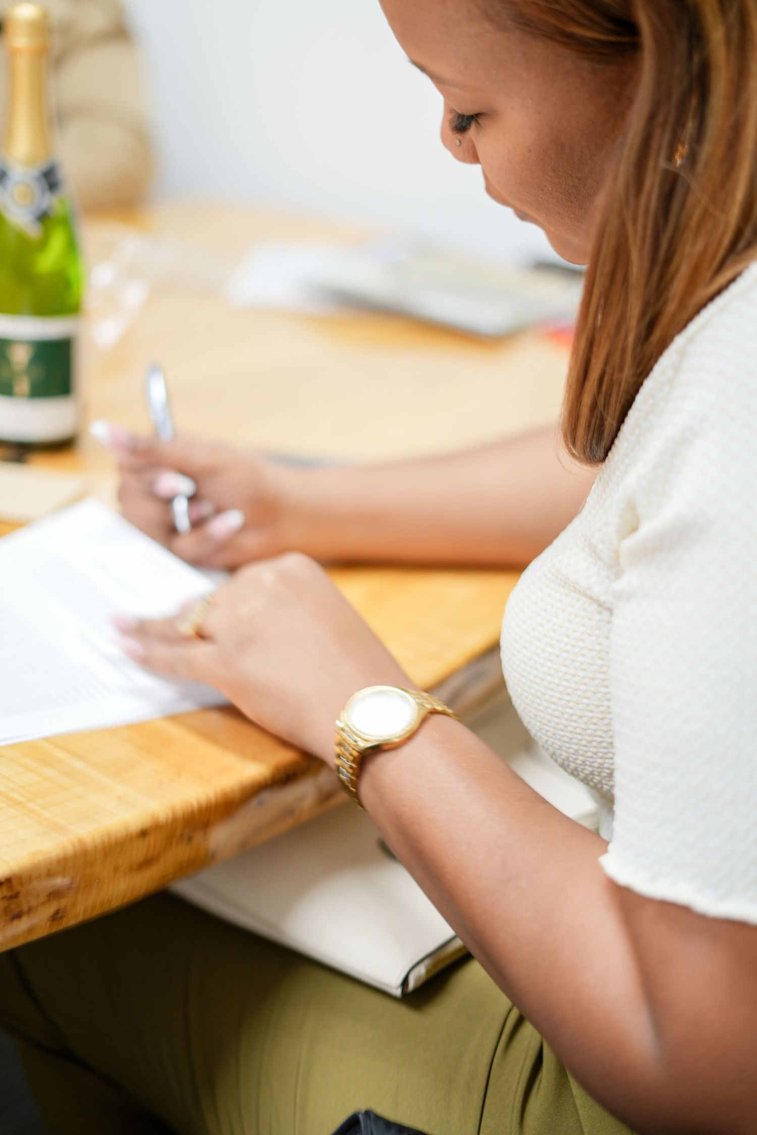 Person writing at wooden desk with champagne bottle nearby.