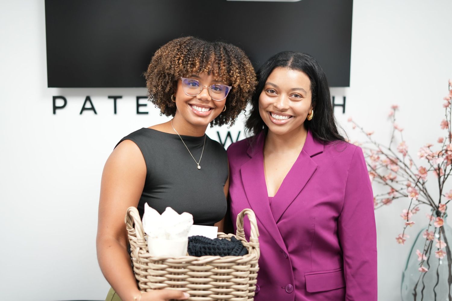 Two professionals smile while holding a gift basket in an office setting.