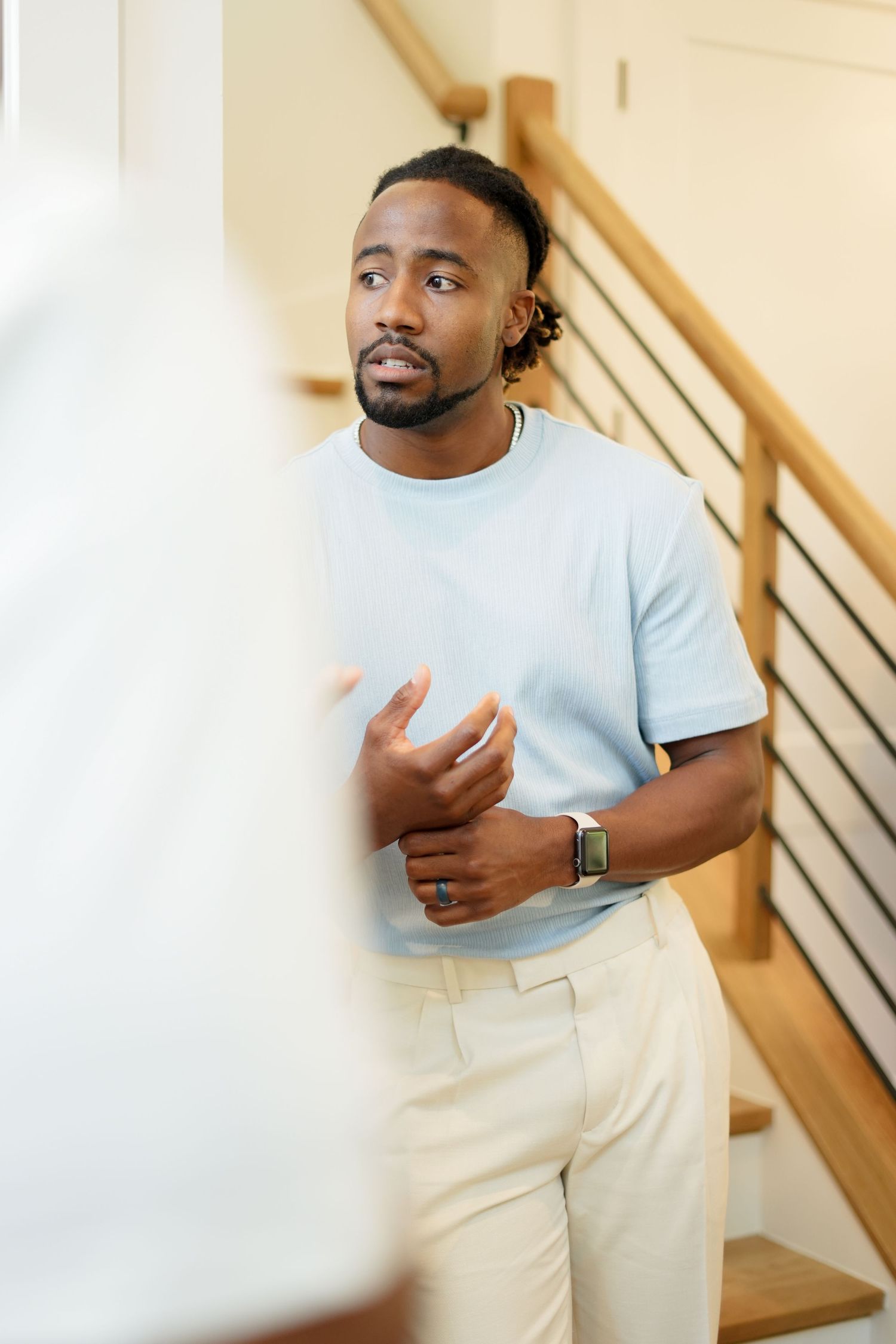 Professional portrait of person in light blue t-shirt and khakis standing near wooden staircase.