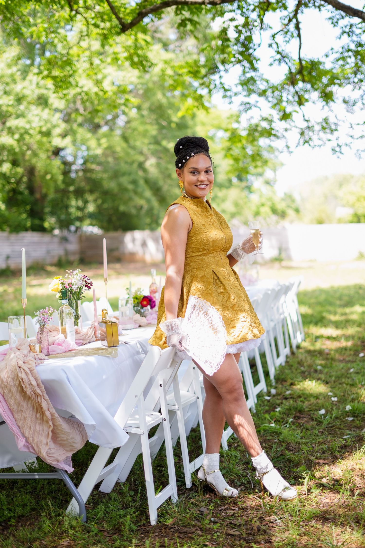 Person in gold dress poses by elegant outdoor table setting with white chairs and floral arrangements.