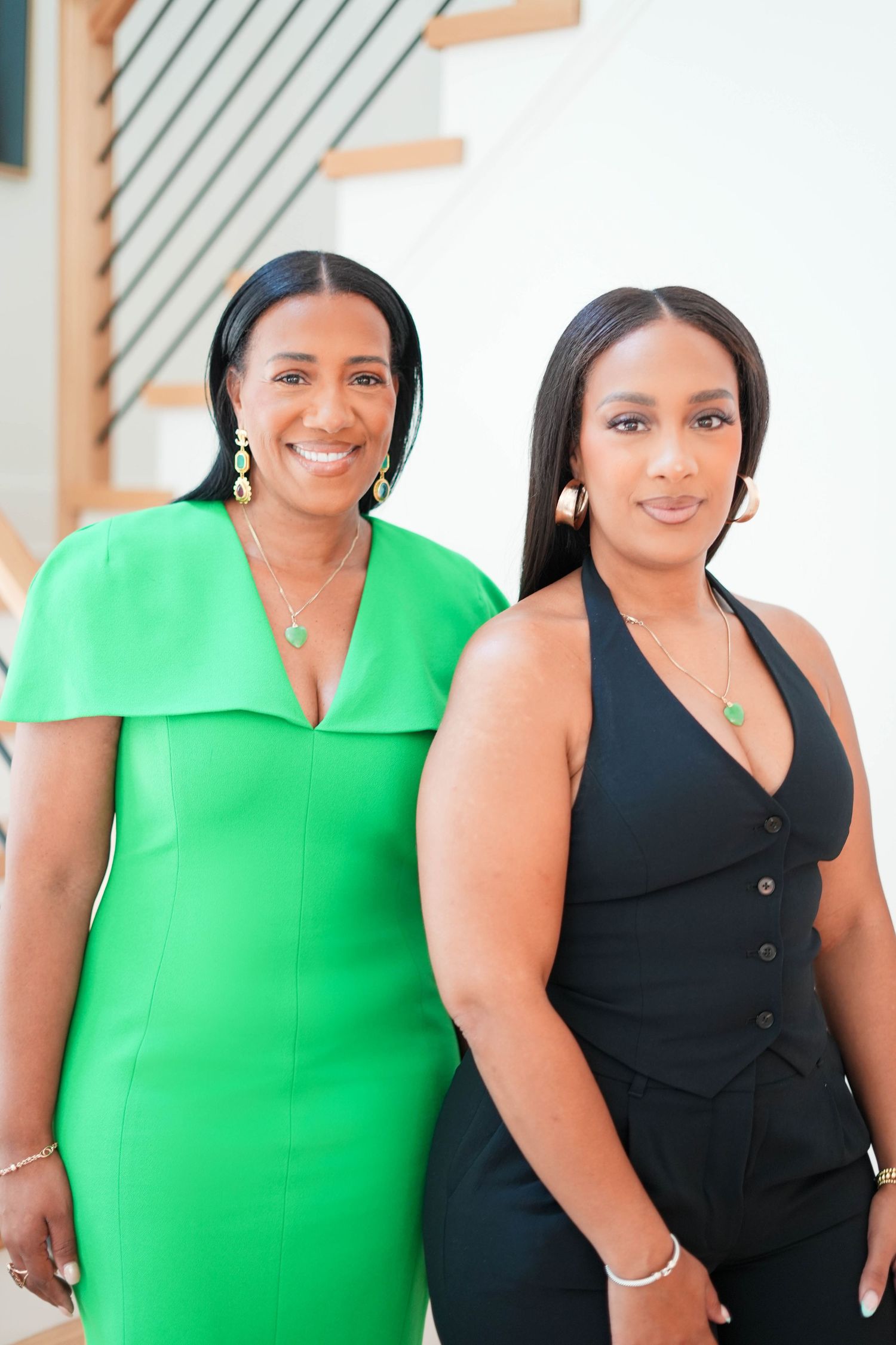 Two women stand together wearing elegant outfits - one in a bright green dress and one in a black ensemble.