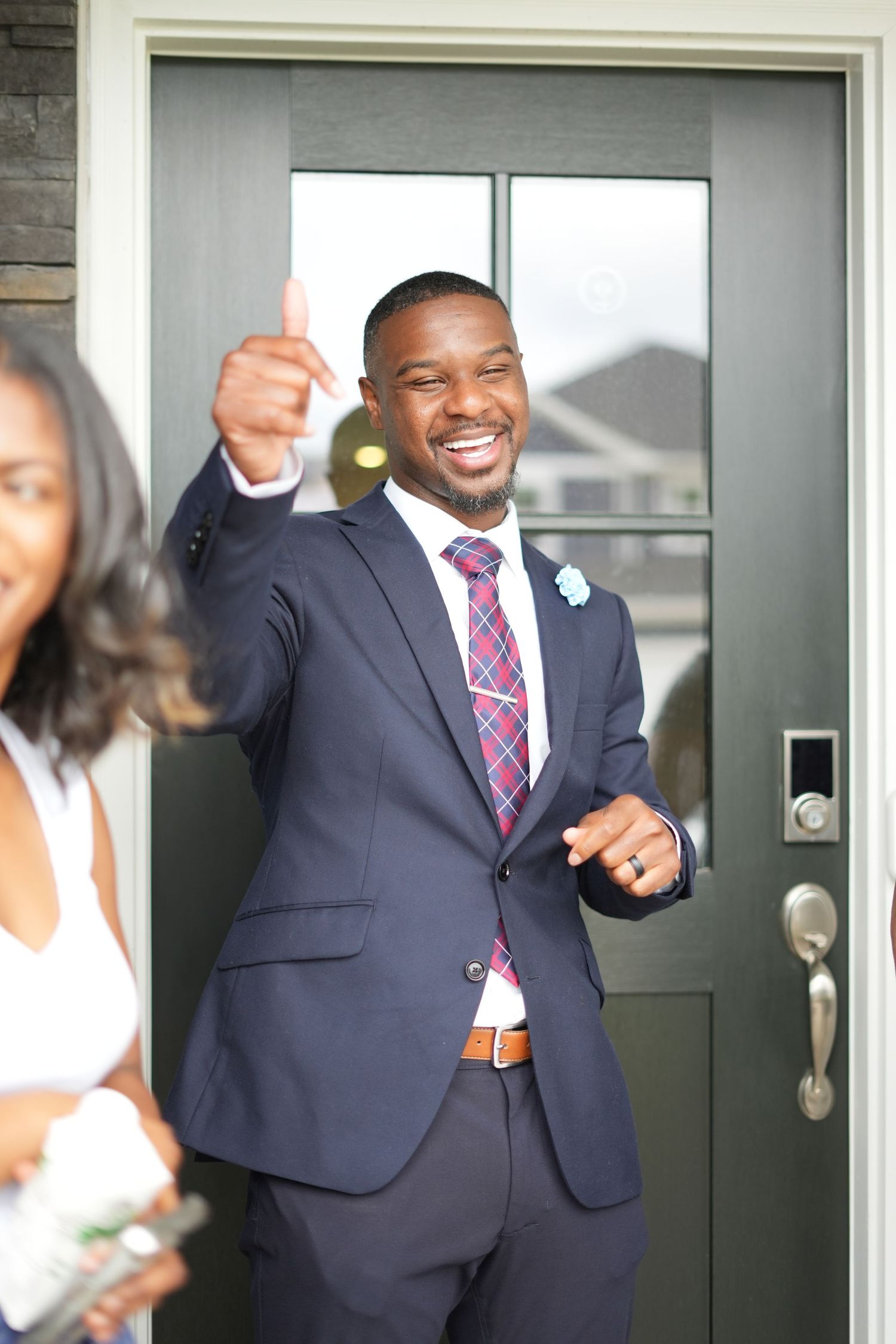 Wedding party member in navy suit and purple tie gives enthusiastic thumbs up while standing in doorway.