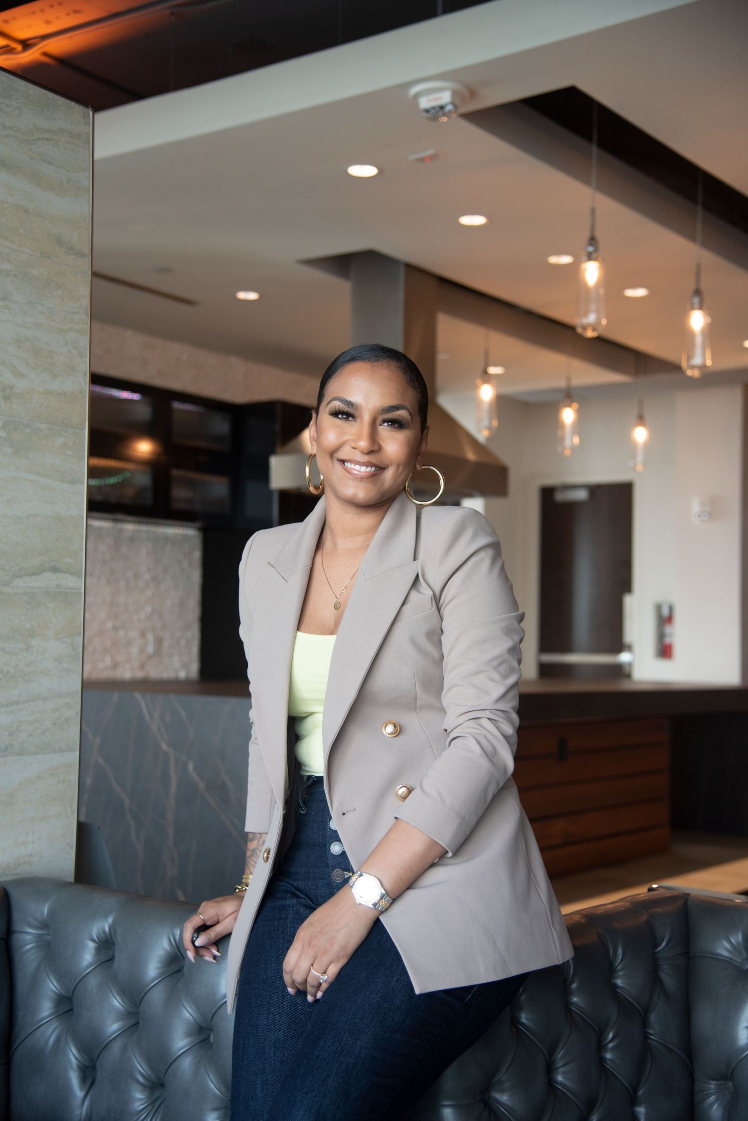 Professional in beige blazer and jeans poses confidently in modern restaurant setting.