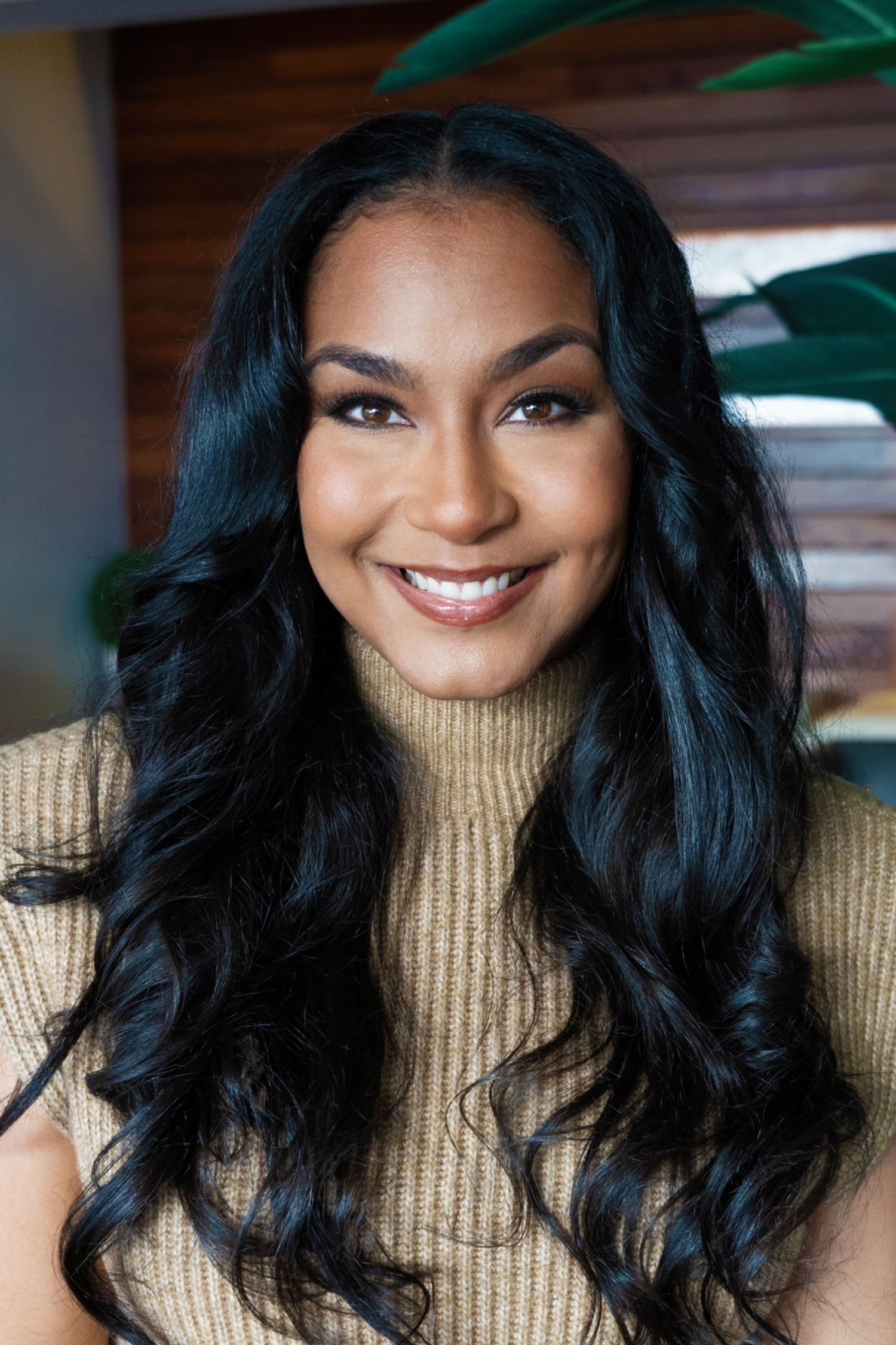 Woman in beige turtleneck sweater with flowing dark hair poses near indoor plant.