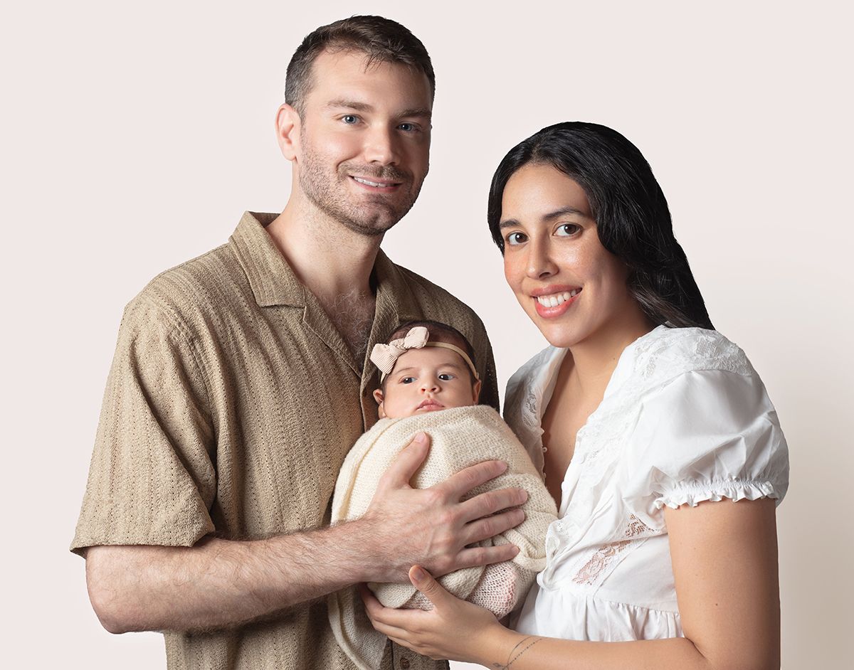 Happy young family portrait with parents holding their newborn baby girl with a pink bow on a neutral background.