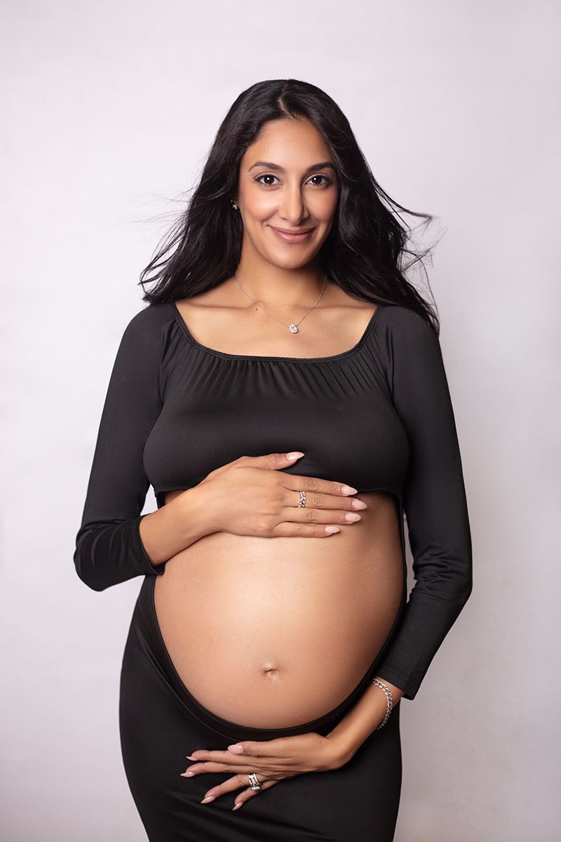 Smiling pregnant woman in black dress cradling her bare baby bump against a white studio background.