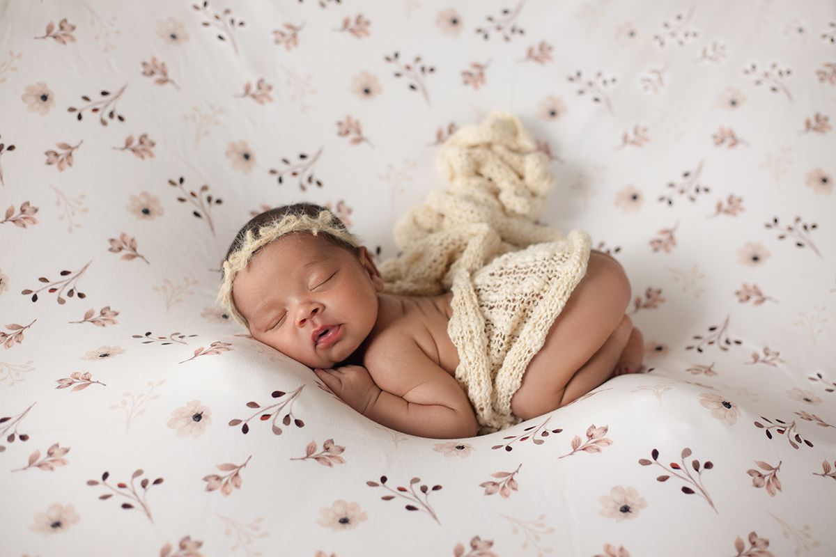 Newborn baby sleeping peacefully on floral blanket wearing cream knit outfit and lace headband.