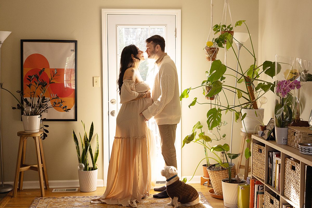 A couple shares a romantic moment by a sunlit door surrounded by lush indoor plants, with a small dog at their feet.