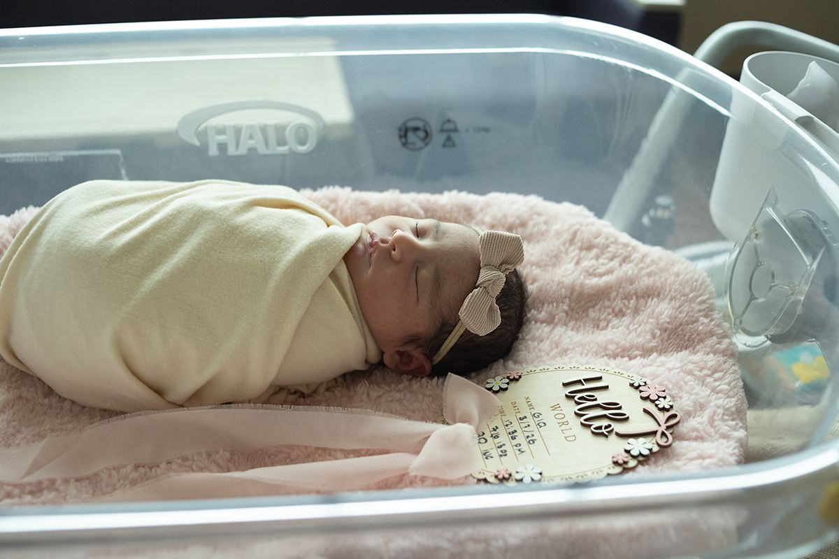 Newborn baby girl swaddled in yellow blanket with bow headband sleeping peacefully in hospital bassinet on pink blanket.