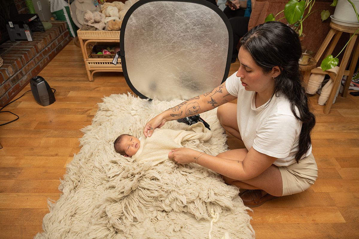 Tattooed woman in white top sits on fluffy rug posing with swaddled newborn baby during professional photo session.