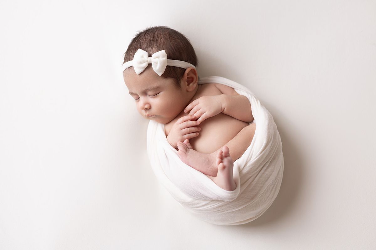 Newborn baby girl swaddled in white wrap with bow headband, sleeping peacefully on white background.