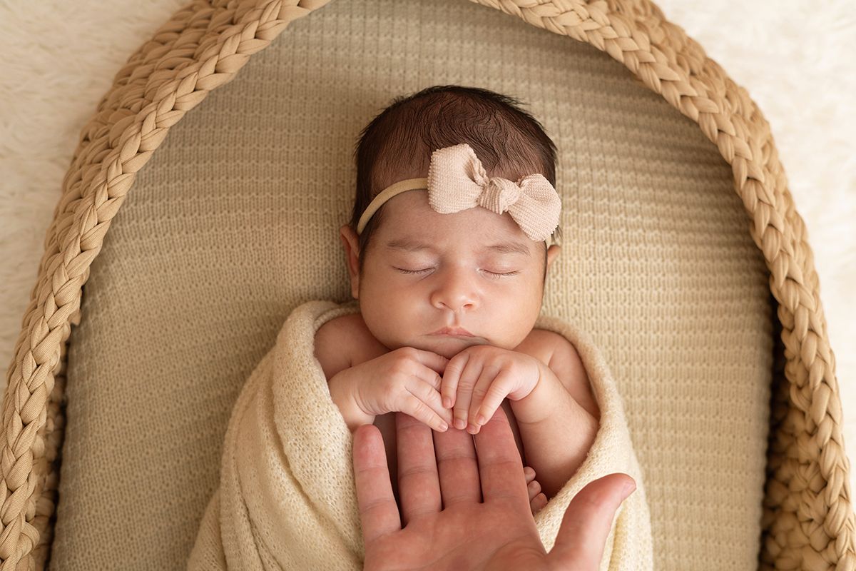 Sleeping newborn baby girl wrapped in cream swaddle with bow headband resting in woven basket with parent's hand.