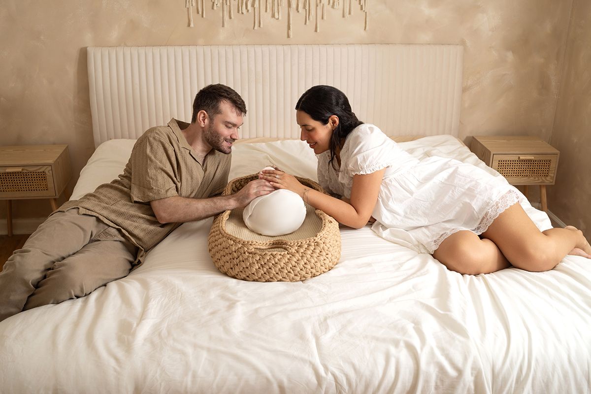 Couple relaxing on a white bed, smiling at each other with a woven hat between them in a cozy bedroom.