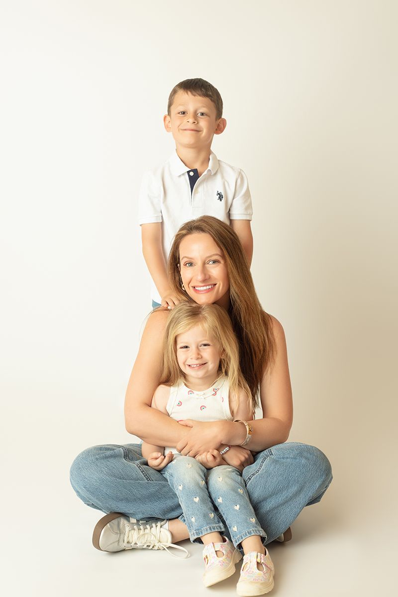 A smiling mother sits cross-legged holding her blonde daughter while her son stands behind her, all wearing jeans.