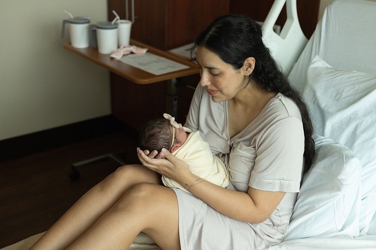 A new mother in a hospital gown sits on a bed, tenderly cradling her newborn baby in her arms.