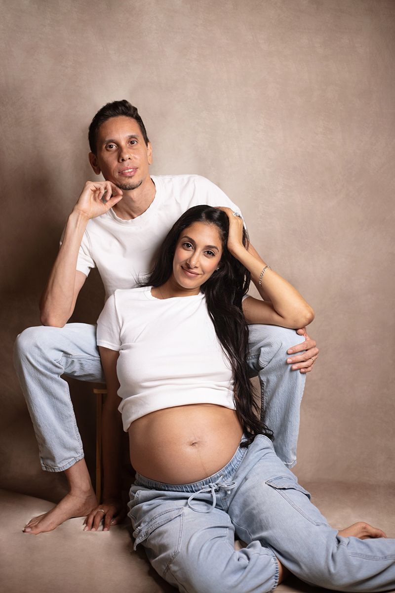 Pregnant couple posing in white tees and jeans, woman sitting on floor showing baby bump, man seated behind her.