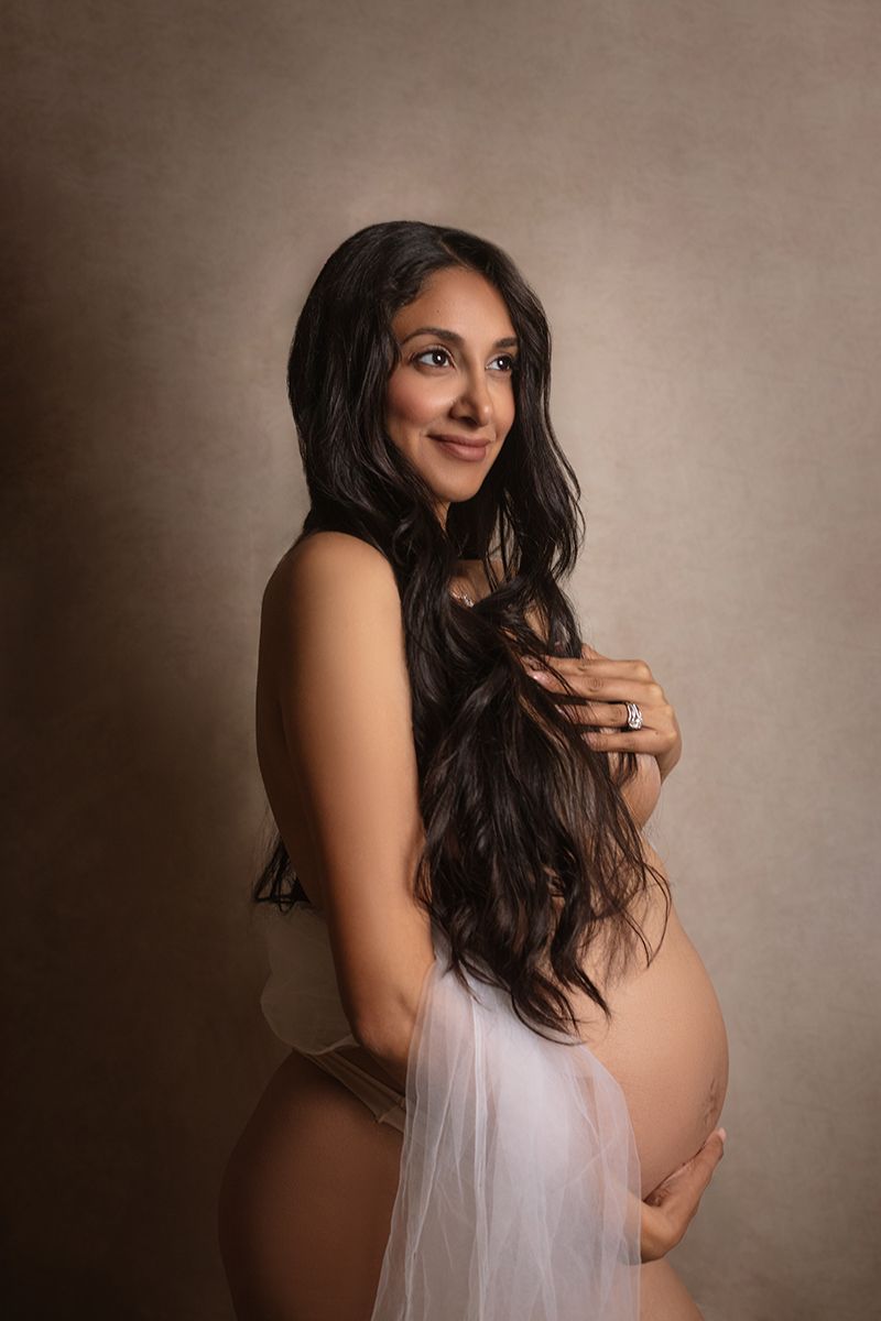 Pregnant woman with long dark hair poses gracefully in sheer white fabric for a studio maternity portrait.