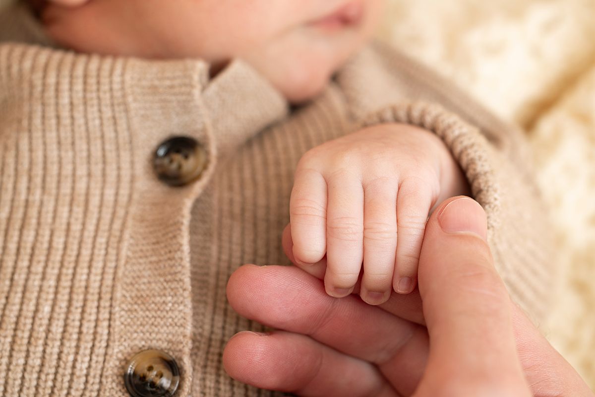 Close-up of a newborn baby gripping an adult's finger, wearing a cozy beige knit cardigan.