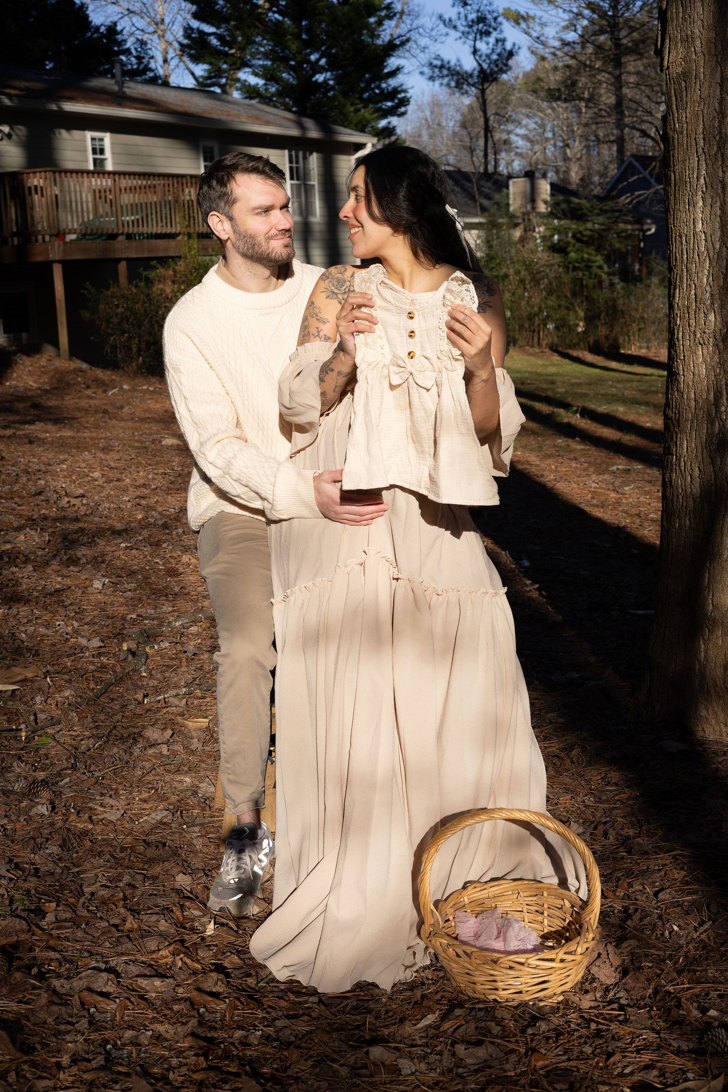 Couple in matching cream outfits standing outdoors near a wicker basket during a maternity photo shoot.