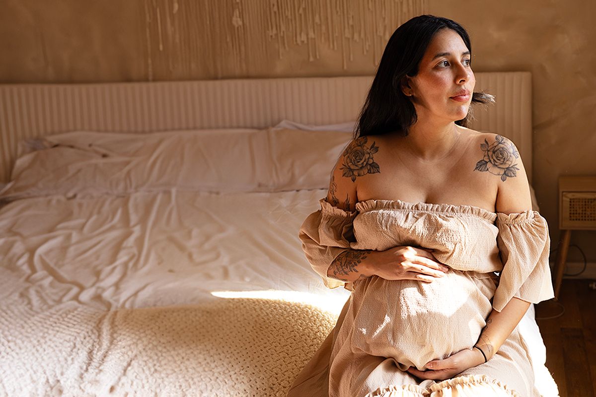 Pregnant woman in blush off-shoulder dress sitting by bed, cradling belly with tattoo on shoulder.