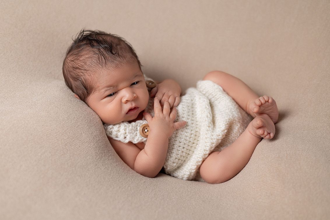 Newborn baby sleeping peacefully in a cream knit romper on a beige backdrop, curled in a natural pose.