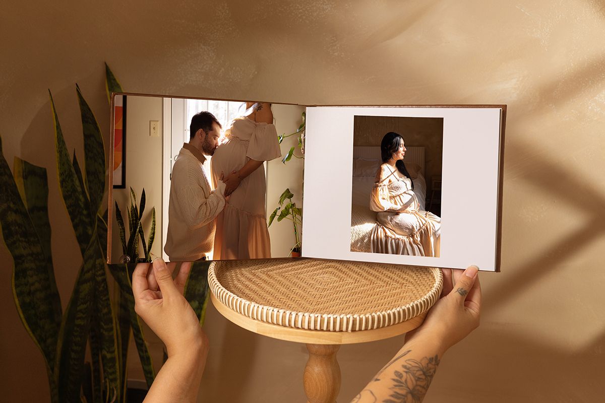 Hands holding an open photo book displaying a couple portrait and a woman in a white dress, set against a warm beige background.