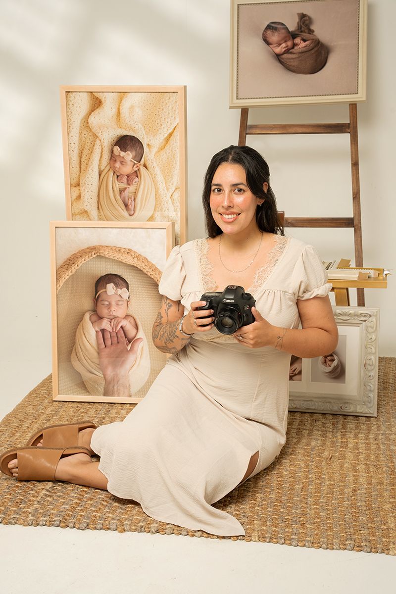 A smiling woman in a cream dress sits on a woven rug holding a camera, surrounded by newborn photography prints.