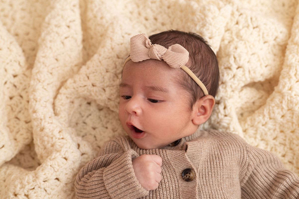 Newborn baby girl in tan knit cardigan and bow headband sleeping on cream crochet blanket.