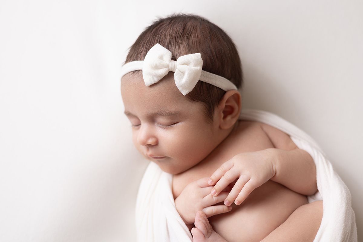 Sleeping newborn baby girl wearing a white bow headband, wrapped in a soft white cloth on a white background.