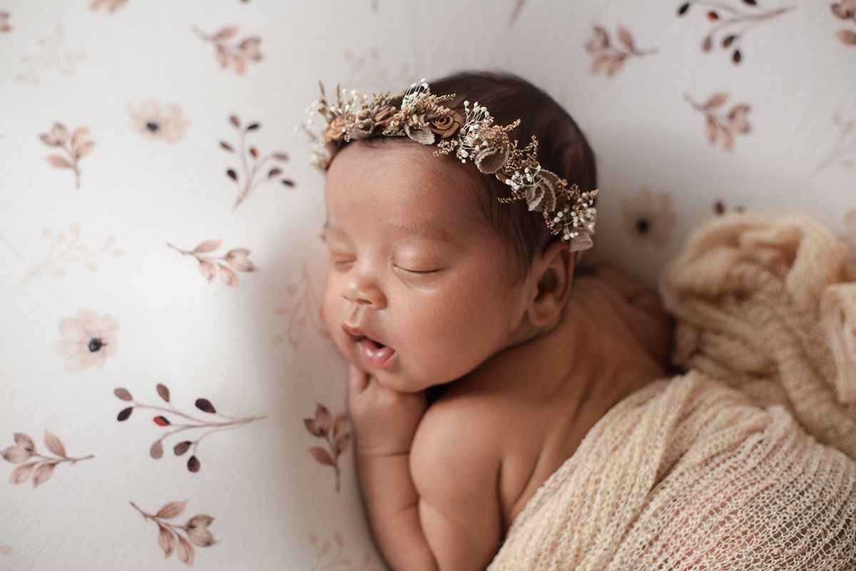 Sleeping newborn baby wearing a delicate floral headband, wrapped in a beige knit blanket on a floral background.