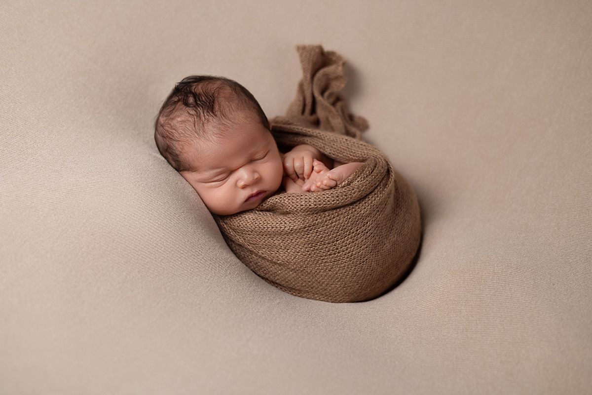 Newborn baby peacefully sleeping wrapped in a soft brown knit swaddle on a beige backdrop.