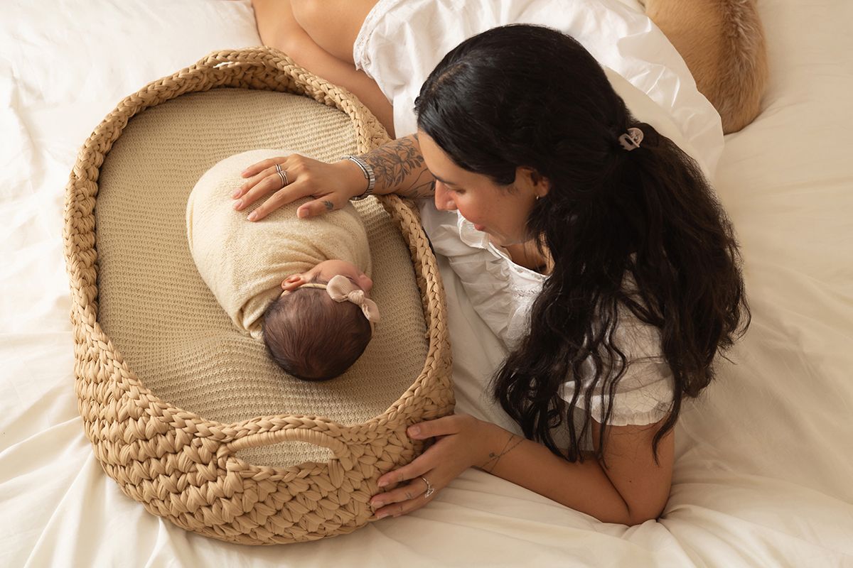 Mother gazing lovingly at her newborn baby resting in a woven Moses basket on a white bed.
