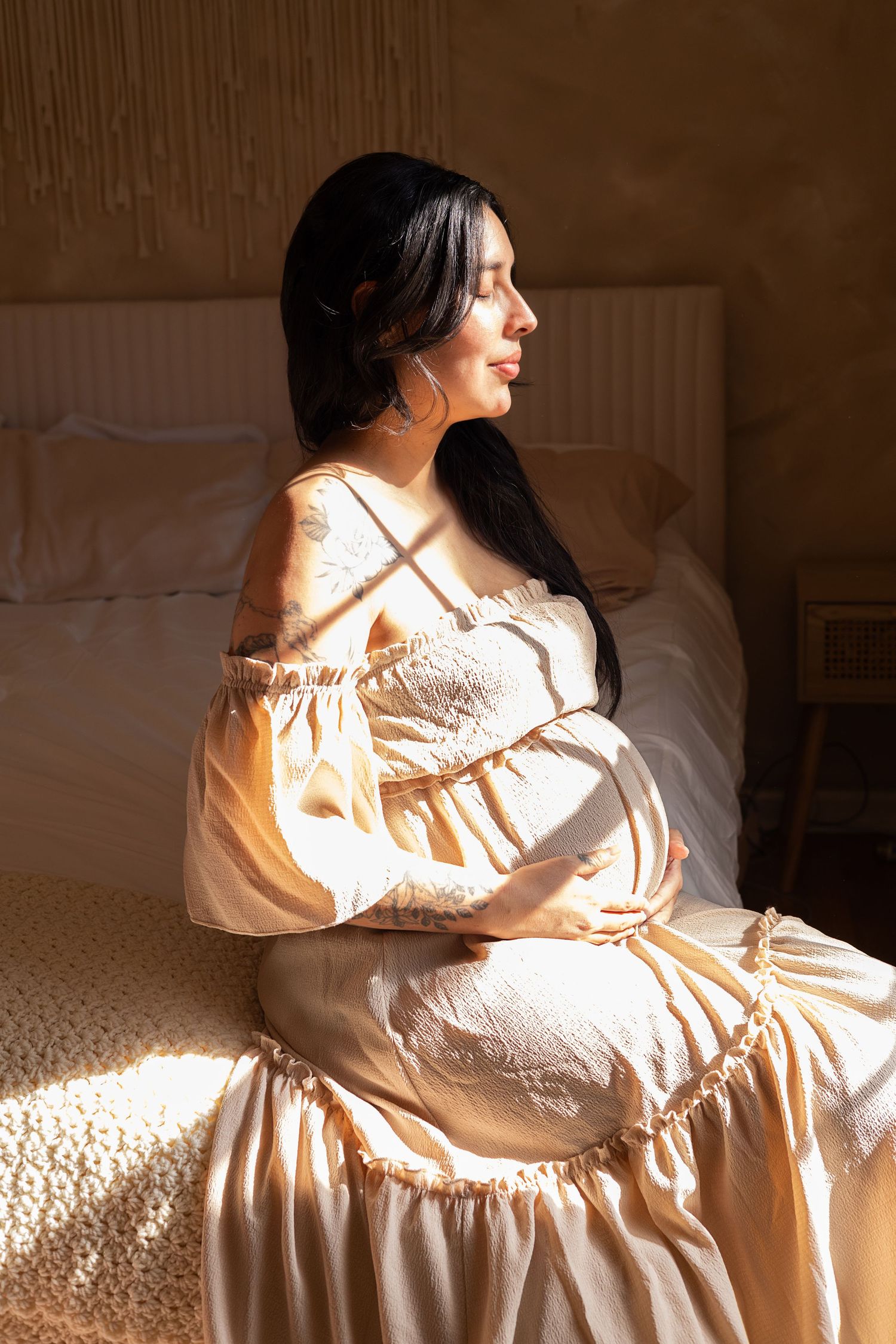 Pregnant woman in a cream boho dress sitting on a bed, cradling her baby bump in warm sunlight.