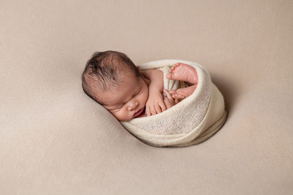 Newborn baby swaddled in cream wrap, peacefully sleeping in a bowl on a beige backdrop.