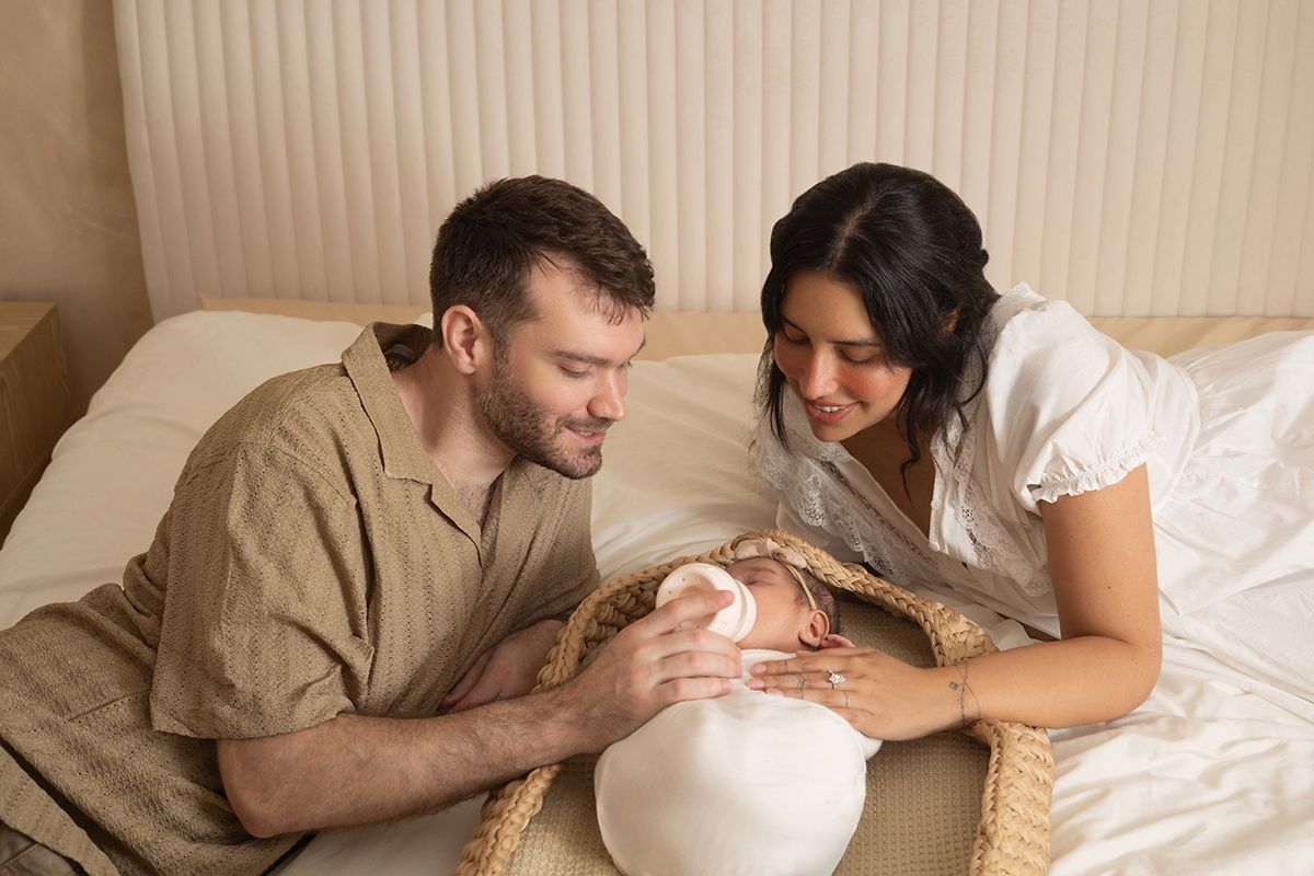 A couple lovingly gazes at their newborn baby while relaxing together on a bed in a soft, neutral-toned room.