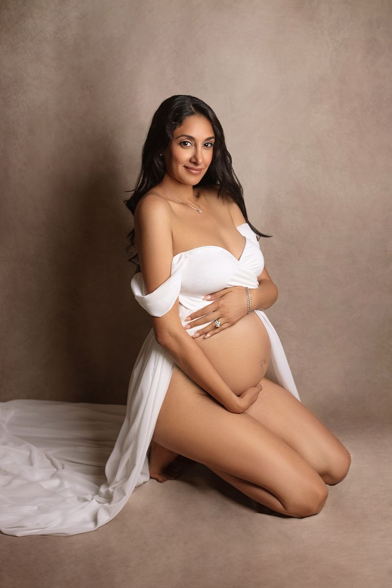 Pregnant woman in white off-shoulder draped gown kneeling gracefully in a professional maternity studio portrait.