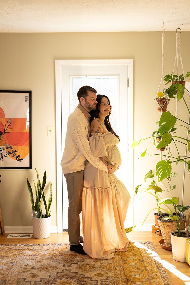 A couple shares a tender embrace in a sunlit boho home, the woman wearing a flowing peach maternity gown surrounded by lush plants.