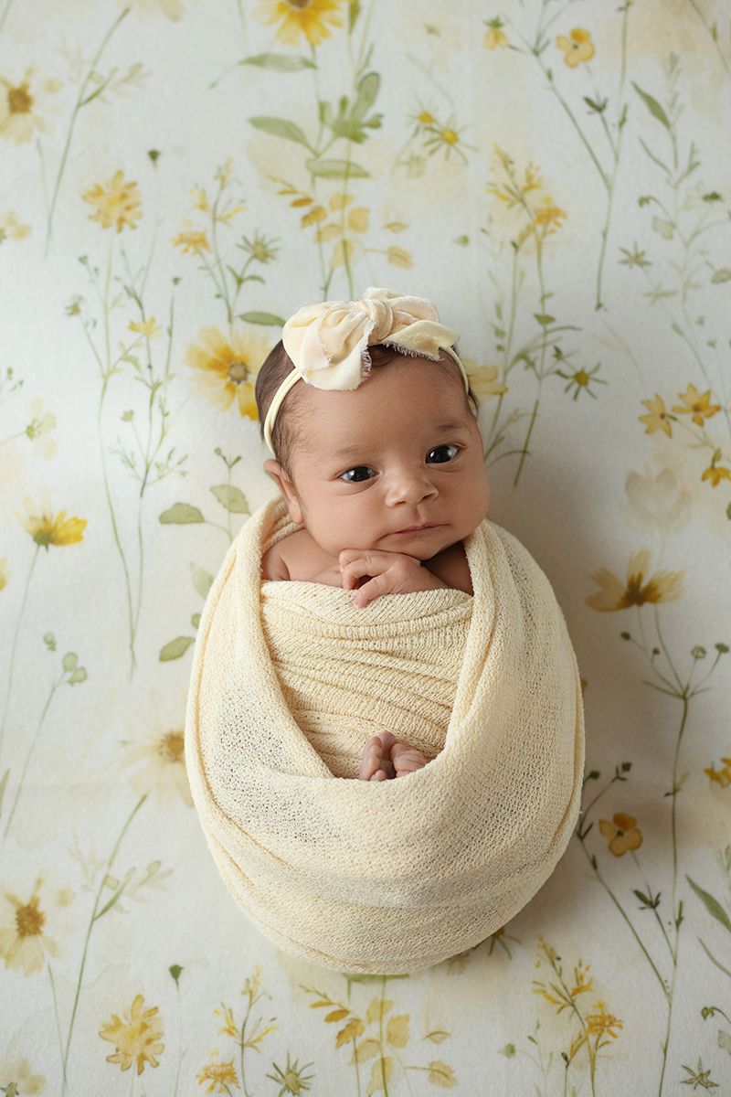 Newborn baby girl swaddled in cream wrap with floral headband, posed on yellow floral backdrop.