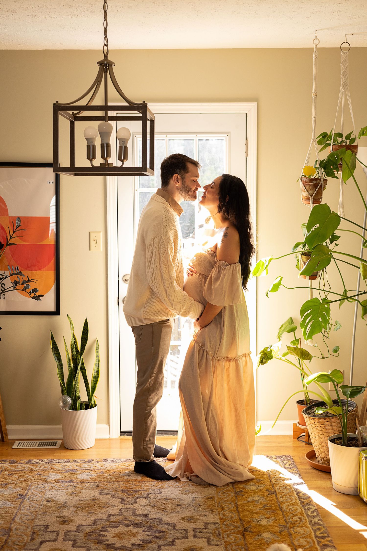 Expectant couple nearly kissing in a warm sunlit entryway surrounded by tropical houseplants and boho decor.
