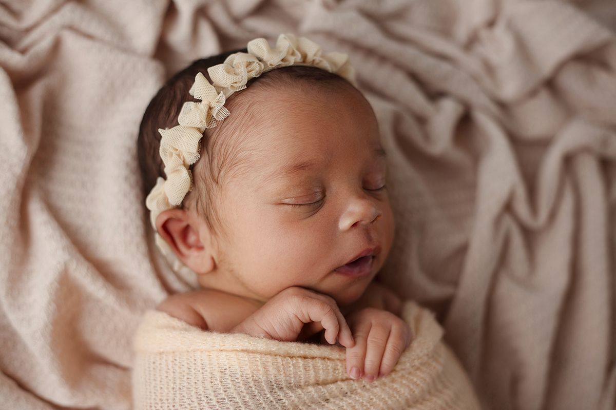 Sleeping newborn baby girl swaddled in cream knit wrap with floral headband on soft pink backdrop.