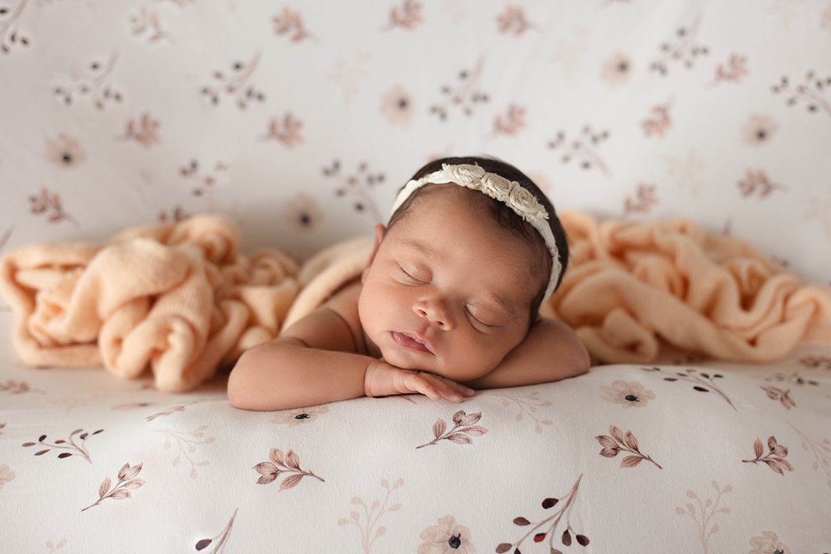 Sleeping newborn baby girl wearing a delicate headband, resting on a floral backdrop with a soft peach knit blanket.