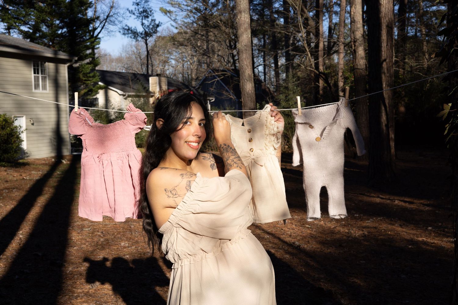 Pregnant woman in a cream dress standing near baby clothes on a clothesline outdoors.
