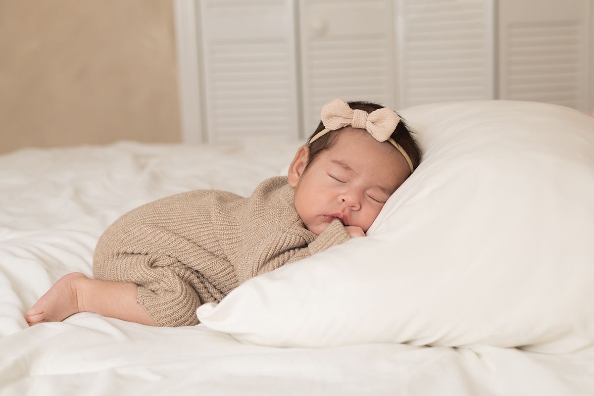 Peaceful newborn baby sleeping on white pillow wearing beige knit romper and pink bow headband.