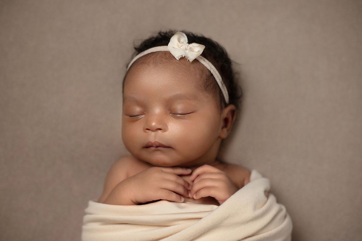 Peaceful newborn baby girl swaddled in cream wrap with white bow headband, sleeping on neutral background.