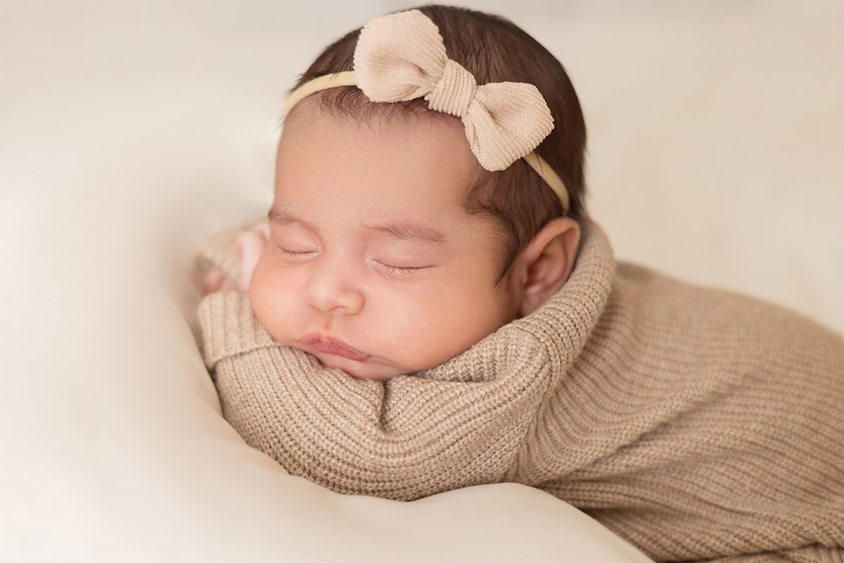 Sleeping newborn baby girl wrapped in a beige knit swaddle wearing a bow headband on a white backdrop.