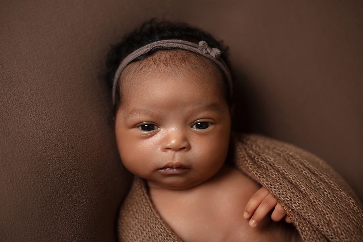 Newborn baby girl wrapped in brown knit swaddle with gray floral headband on brown background.